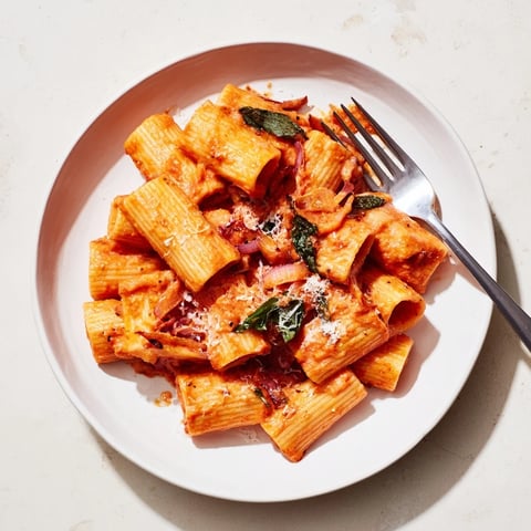 A steaming bowl of spicy rigatoni pasta features creamy tomato sauce clinging to each tube, topped with fresh basil and shaved Parmesan.
