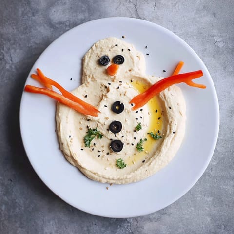 Vibrant melted snowman hummus platter: creamy hummus decorated with carrot, olives, and red pepper for a fun appetizer.